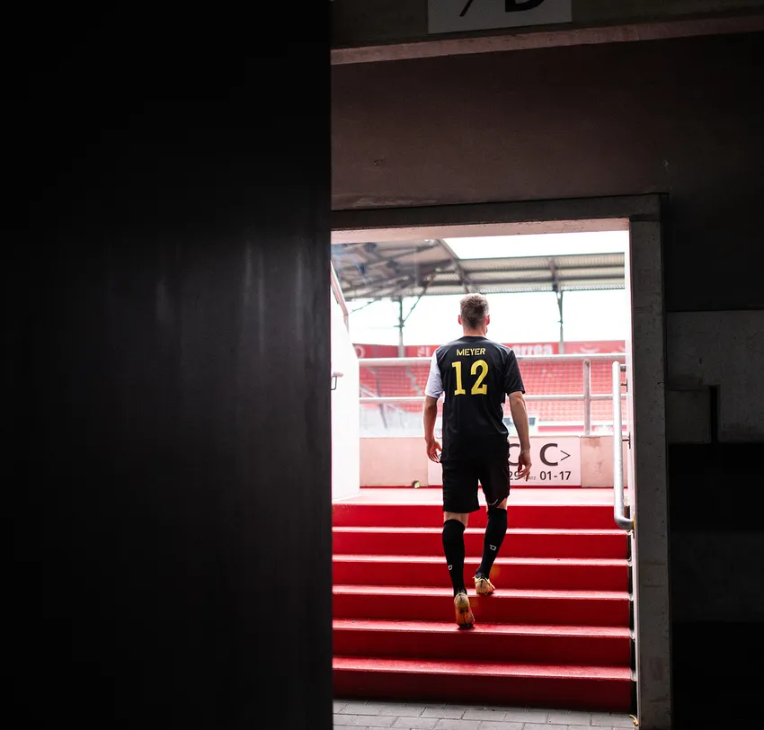 Un futbolista con una camiseta negra con la inscripción 'MIEYER 12' en amarillo sube las escaleras rojas de un estadio.