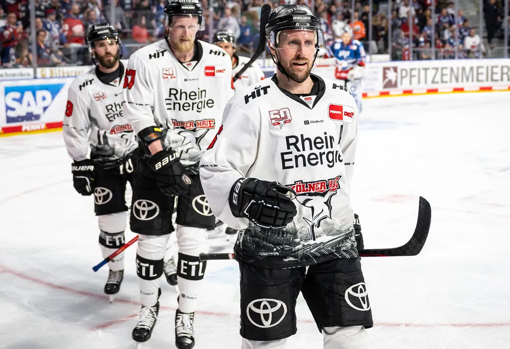 Jugadores de hockey sobre hielo con camisetas blancas y pantalones negros que llevan el logo de un equipo, de pie sobre el hielo.