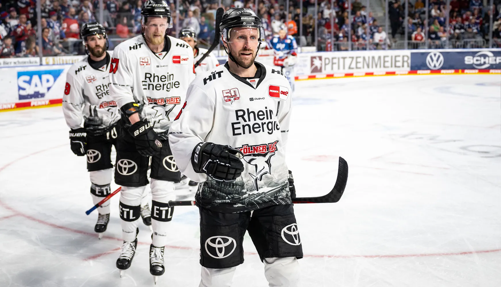 Tres jugadores de hockey sobre hielo en camisetas blancas con diferentes números están de pie sobre el hielo durante un partido.