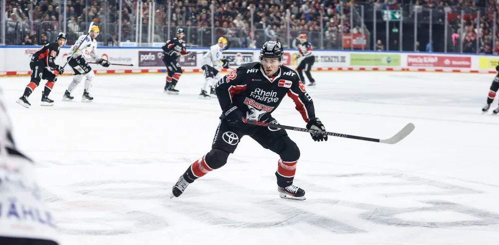 Un jugador de hockey sobre hielo con una camiseta negra con acentos rojos y blancos patinando sobre el hielo con un stick, mientras los espectadores animan en el fondo.