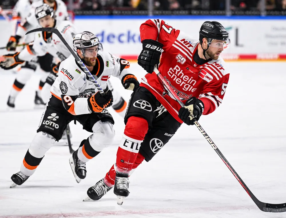 Un jugador de hockey sobre hielo con camiseta roja y pantalones negros patina sobre el hielo mientras es perseguido por otro jugador.