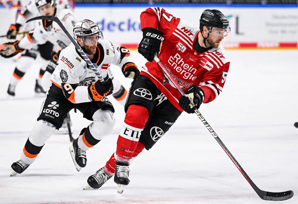 Un jugador de hockey con una camiseta roja patina con el puck sobre el hielo mientras otro jugador con una camiseta blanca lo sigue de cerca.