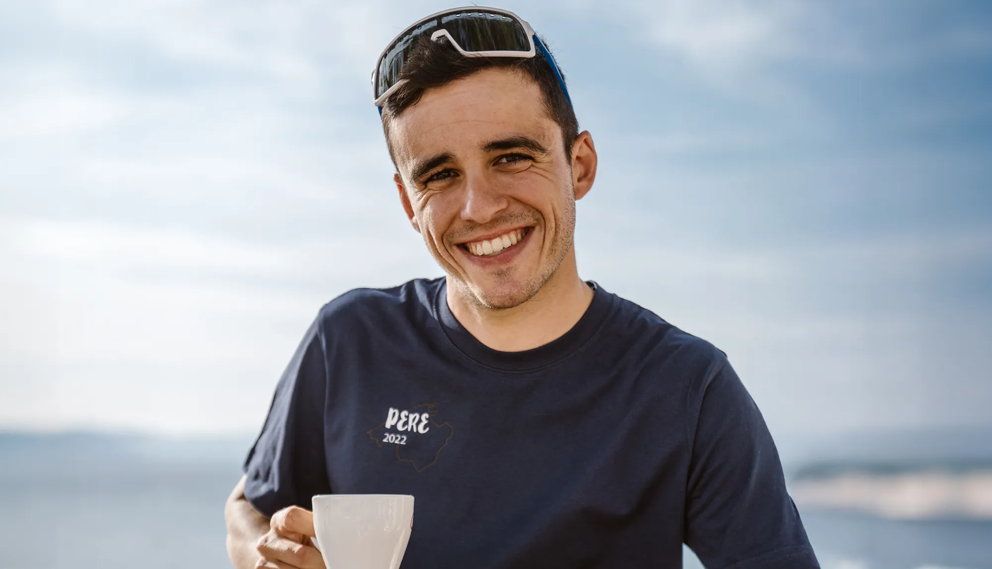 Un joven sonríe con una camiseta azul que presenta el texto 'PERE 2022' mientras sostiene una taza, frente a un cielo despejado.