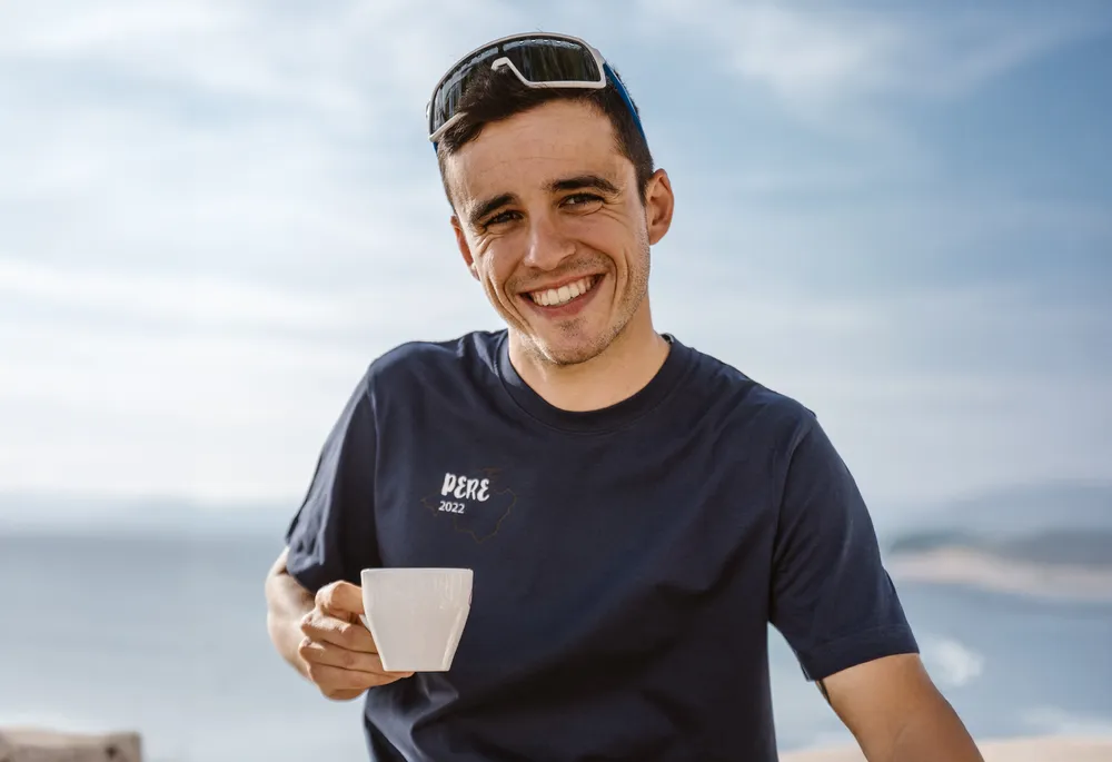 Un hombre sonriente con una camiseta azul sostiene una taza en la mano, con un hermoso paisaje costero de fondo.