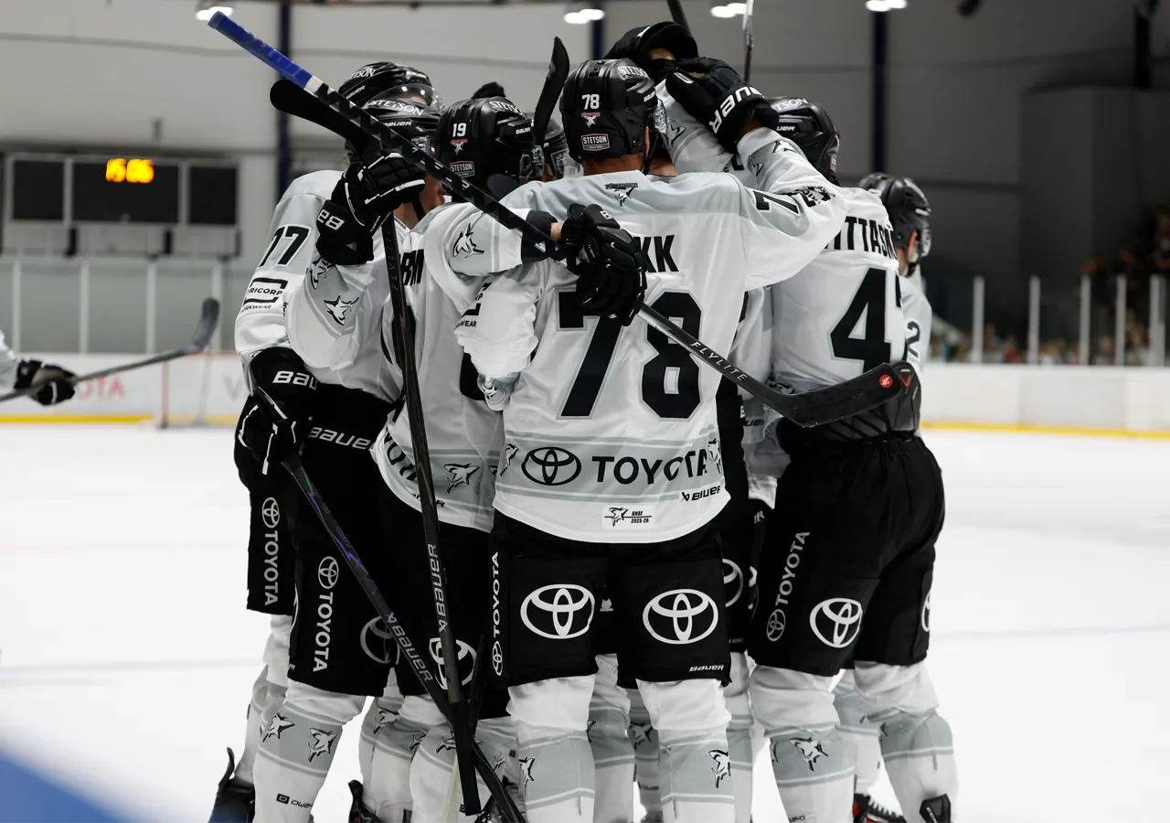 Jugadores de hockey sobre hielo con uniformes blancos con números negros y logos de patrocinadores, abrazándose en la pista de hielo.