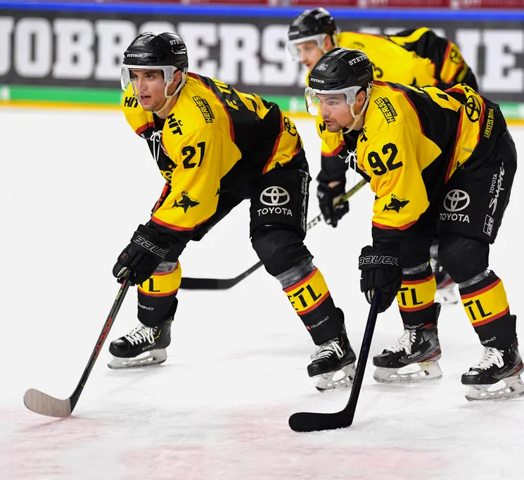 Jugadores de hockey sobre hielo con uniformes amarillos y negros con los números 21 y 92 en la pista, sosteniendo sus palos.