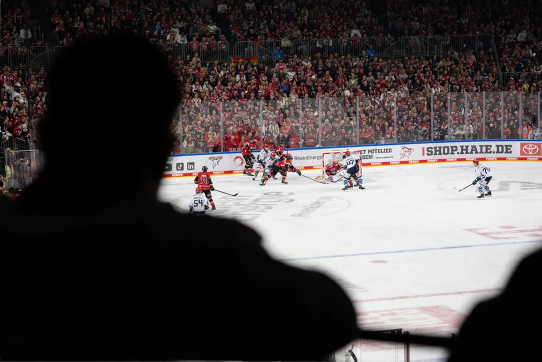 Desde el punto de vista de un espectador, la imagen muestra un partido de hockey sobre hielo con jugadores en camisetas rojas y azules frente a una gran multitud.
