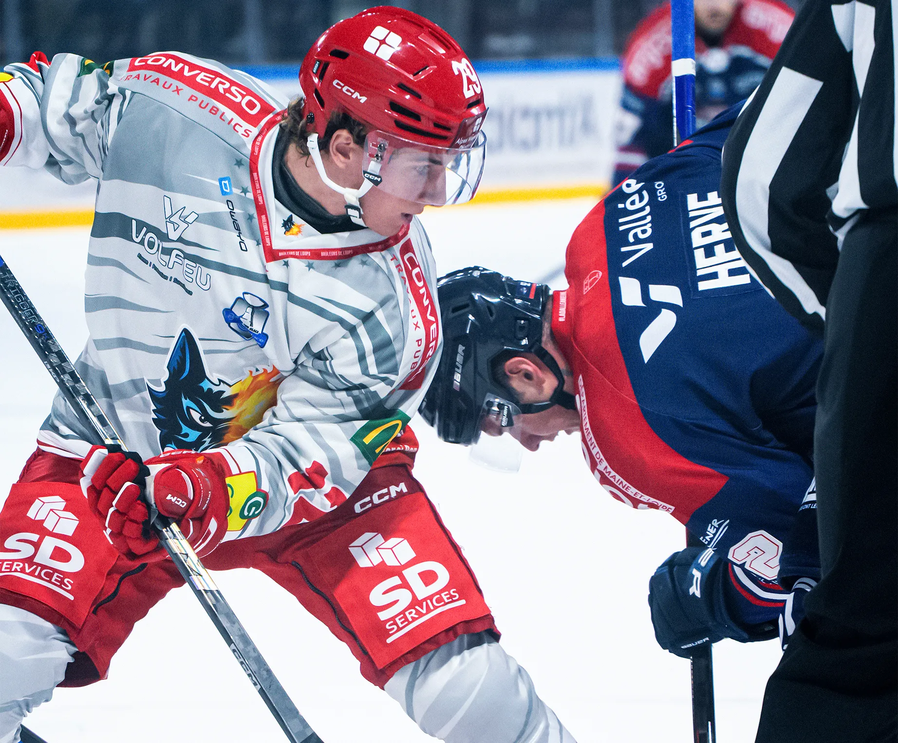 Dos jugadores de hockey sobre hielo se enfrentan, listos para un saque, uno lleva una camiseta gris y roja con un diseño de lobo.