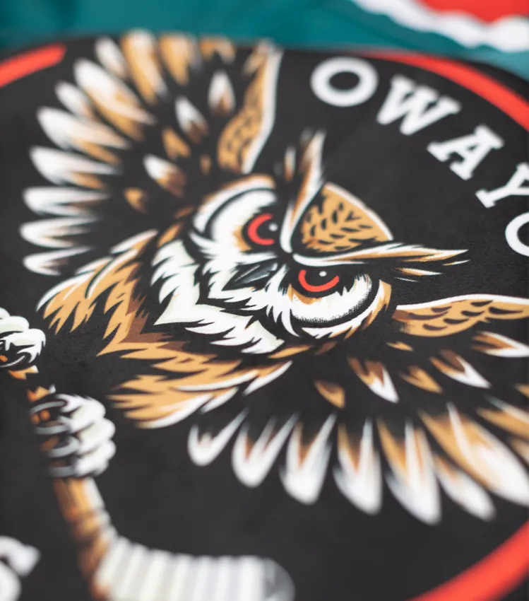 Un primer plano de una camiseta deportiva negra con un logotipo de búho estilizado sosteniendo un palo de hockey, y el texto 'owayo' en letras blancas.