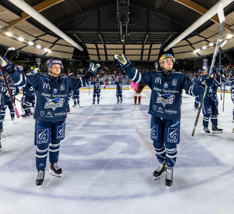 Jugadores de hockey sobre hielo con uniformes azules y logos de patrocinadores celebran en la pista, con el público en el fondo.