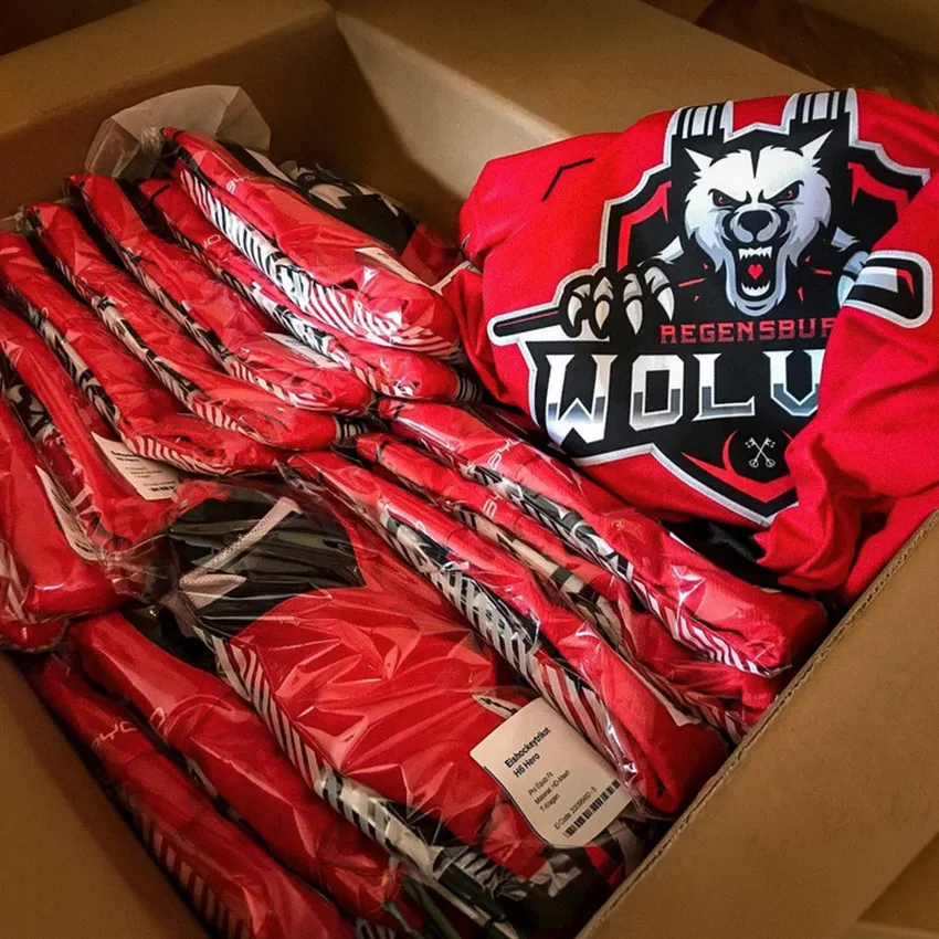 Una caja llena de varias camisetas deportivas rojas envueltas en plástico, incluyendo una camiseta visible con un logo de lobo y el texto 'Regensburg WOLV'.