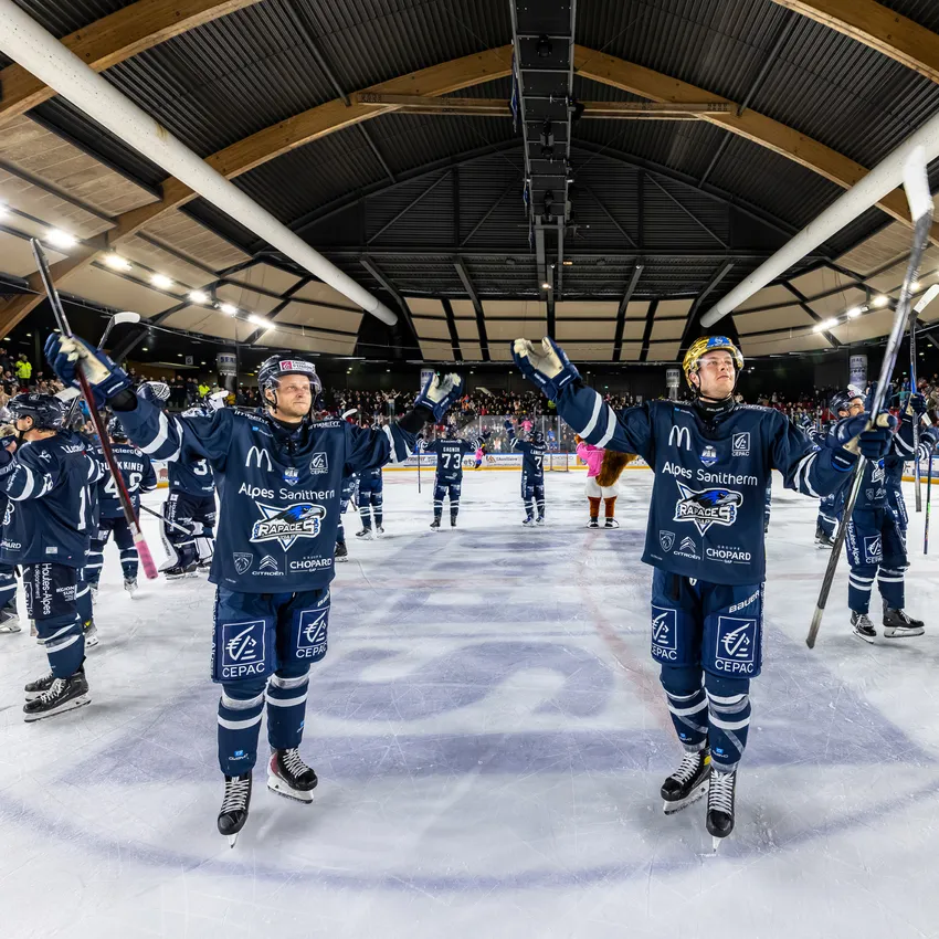 Dos jugadores de hockey sobre hielo con camisetas azules de pie sobre el hielo, levantando sus palos mientras una multitud animada es visible al fondo.