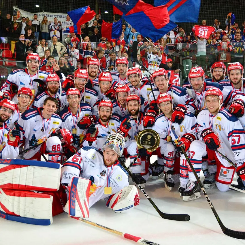 Un grupo de jugadores de hockey sobre hielo con camisetas blancas con acentos rojos y azules posa orgullosamente con medallas y un trofeo sobre el hielo.