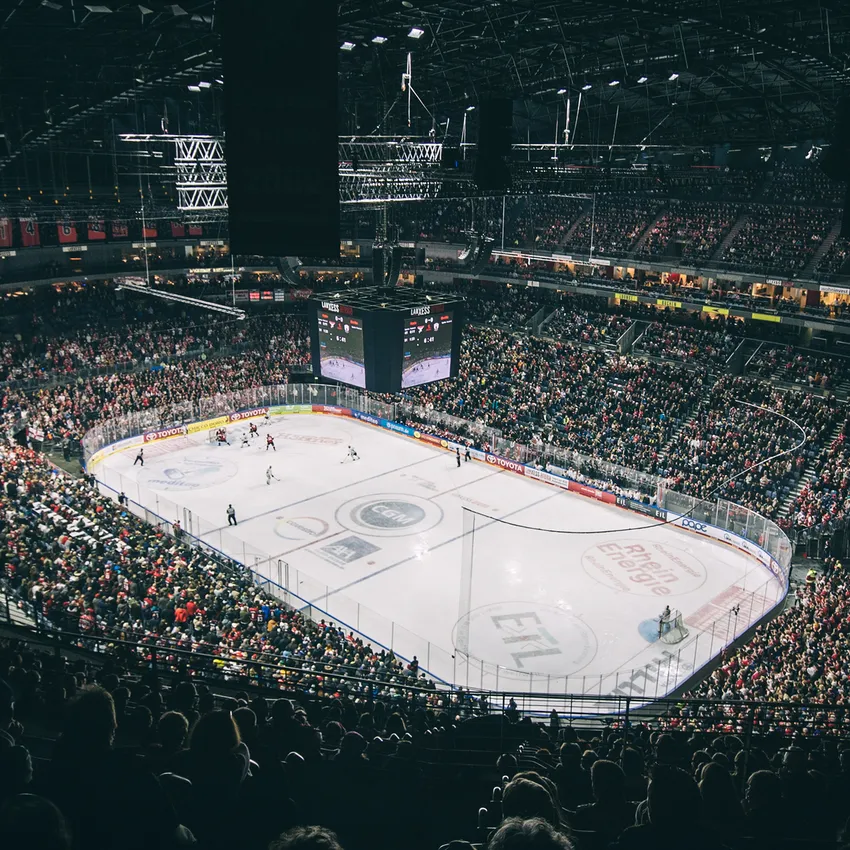 Una arena de hockey sobre hielo llena de aficionados animando, con una pista visible y una gran pantalla en el fondo.