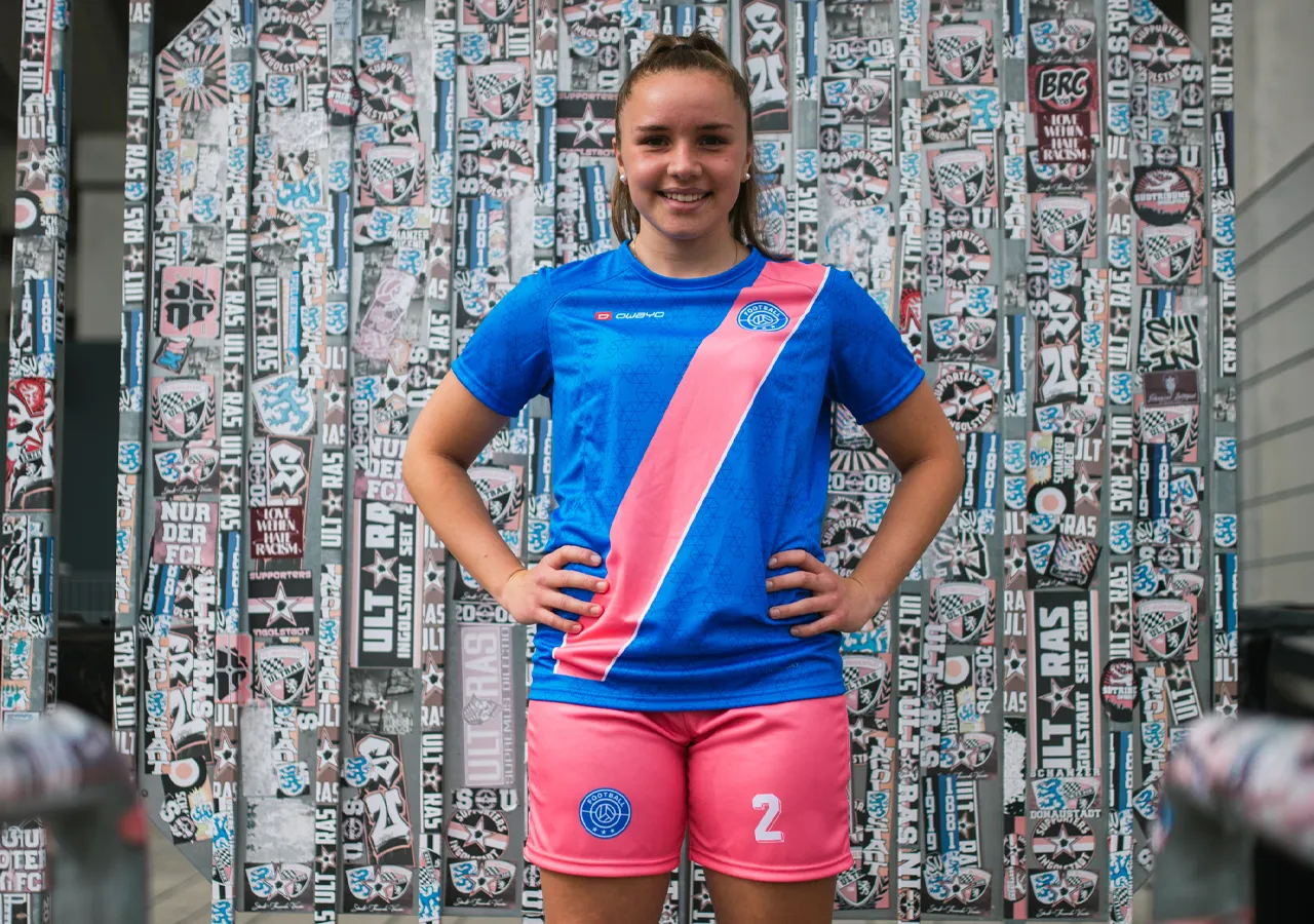 Una joven lleva una camiseta de fútbol azul con una franja diagonal rosa y pantalones cortos rosas con el número 2, frente a una pared cubierta de gráficos y pegatinas.