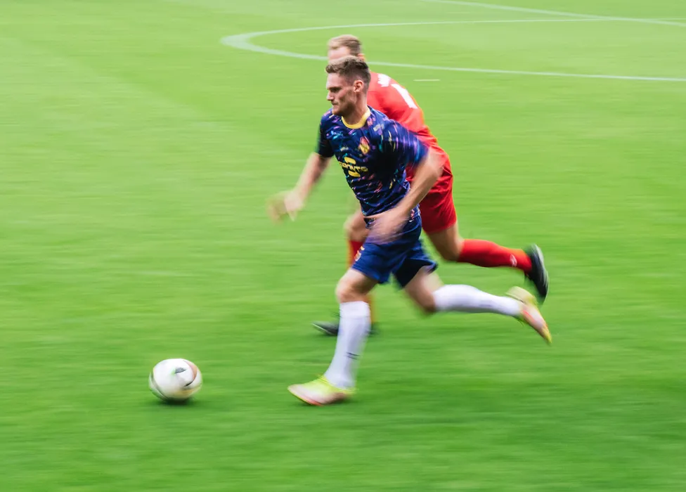 Un jugador de fútbol con una camiseta azul de patrones coloridos corre rápidamente por el campo verde mientras otro jugador lo sigue de cerca.