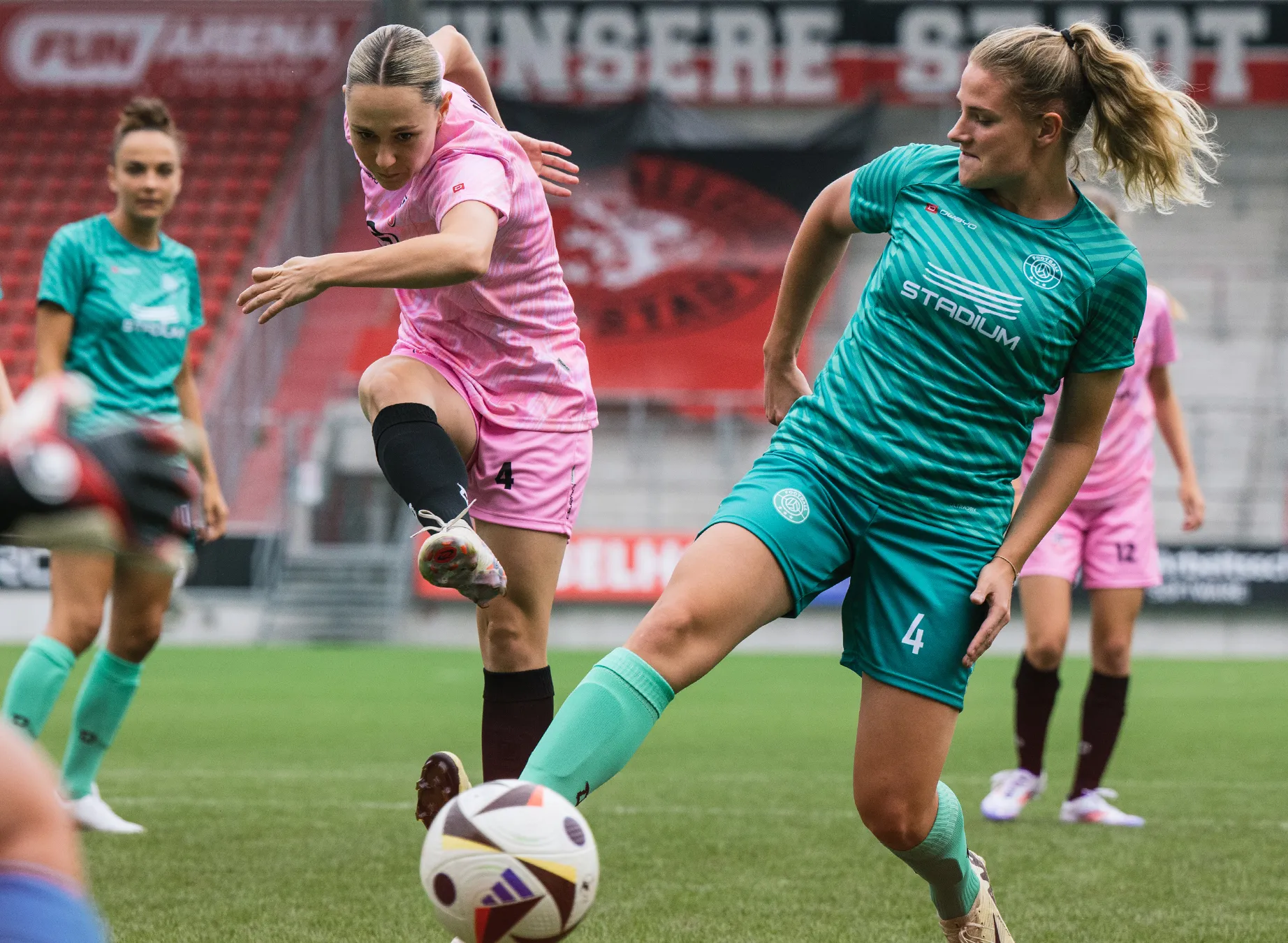 Dos jugadoras de fútbol con camisetas brillantes, una en rosa y la otra en verde, están jugando en un campo de césped.