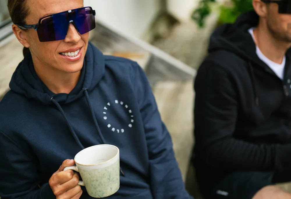 Una persona con una sudadera azul con un logo sostiene una taza mientras está sentada en un banco de madera.
