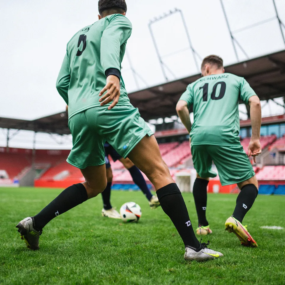 Dos futbolistas en camisetas verdes y calcetas negras jugando activamente en un campo de césped en un estadio.