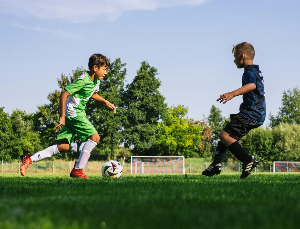 Dos niños juegan al fútbol en un campo verde, uno lleva una camiseta verde y el otro una negra, mientras persiguen el balón.