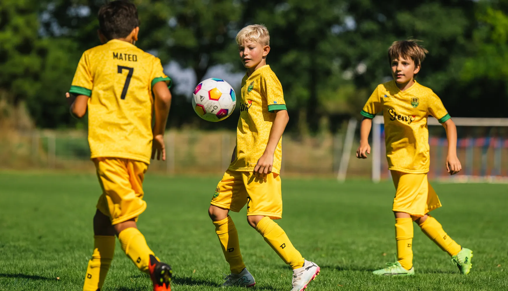 Tres niños con camisetas de fútbol amarillas juegan en un campo de hierba con un balón de fútbol colorido.