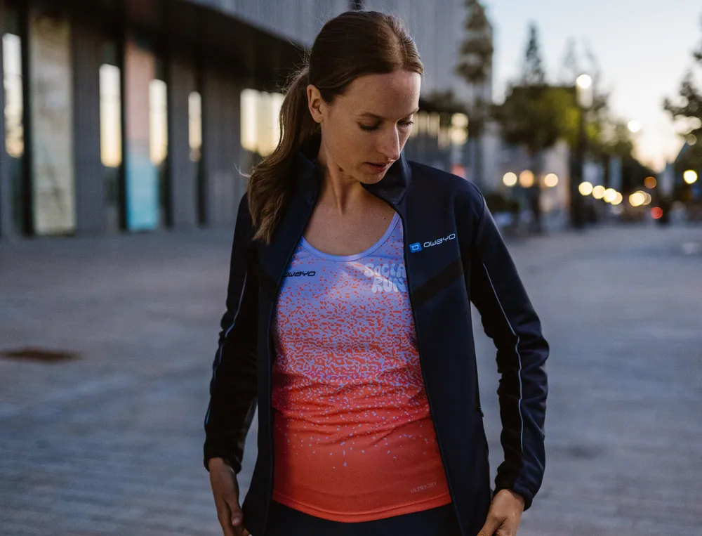 Una mujer con ropa deportiva, que consiste en una chaqueta ligera y una parte superior colorida, está de pie en un entorno urbano, mirando pensativamente su atuendo.