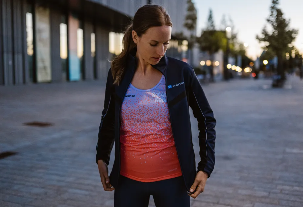 Una mujer atlética lleva una chaqueta azul oscura sobre una camiseta sin mangas de degradado azul y naranja mientras está en un entorno urbano.