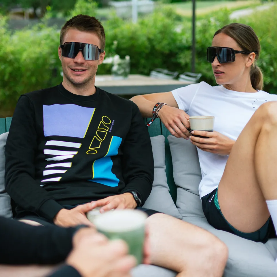 Dos personas están sentadas en un sofá al aire libre, el hombre lleva un sudadera negra con un diseño colorido y la mujer una camiseta blanca, ambos sosteniendo bebidas.