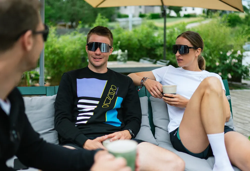 Tres personas en un sofá, dos vistiendo ropa deportiva casual y gafas de sol, mientras se relajan y charlan sosteniendo bebidas.