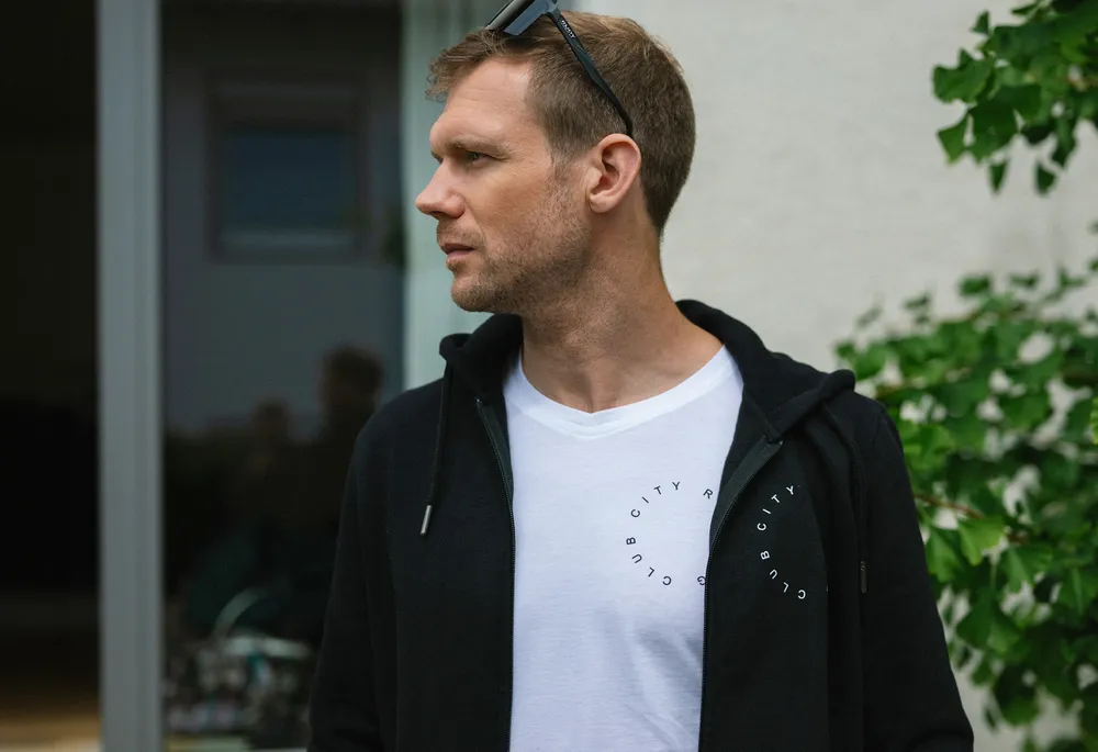 Un hombre está de pie de lado con una sudadera negra y una camiseta blanca con un estampado circular.