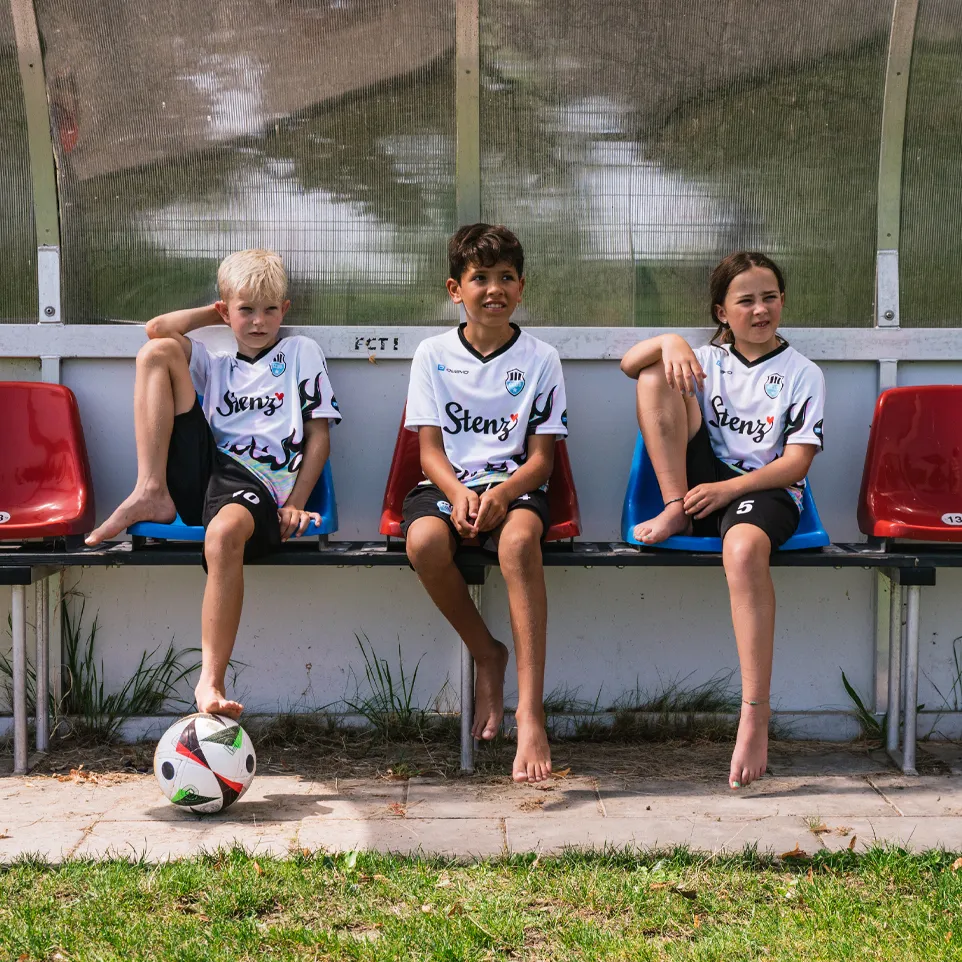 Tres niños con camisetas de fútbol y pantalones cortos están sentados en un banco al lado del campo, con un balón de fútbol frente a ellos.