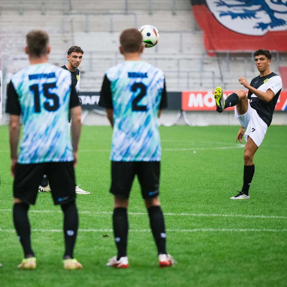 Tres futbolistas con camisetas coloridas están en el campo mirando a otro jugador que está haciendo malabares con un balón.