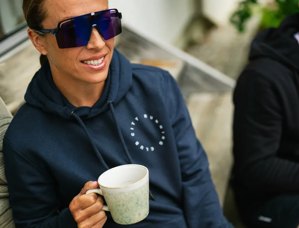Una persona con una sudadera azul marino con un logo sostiene una taza de café y lleva grandes gafas de sol.