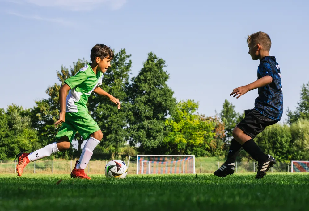 Dos niños están jugando al fútbol en un campo verde, uno lleva una camiseta verde con acentos blancos y el otro una camiseta azul, mientras persiguen un balón.