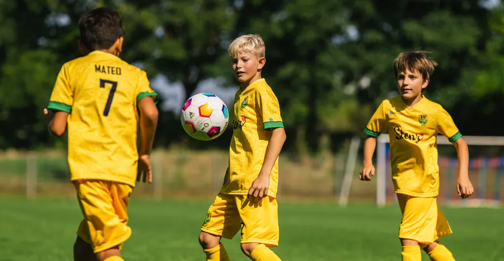 Tres niños en camisetas de fútbol amarillas juegan en un campo verde, mientras uno de ellos controla un balón de fútbol colorido.