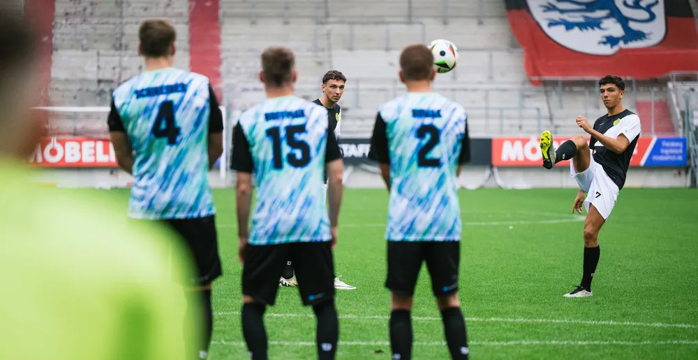 Un jugador de fútbol está realizando un tiro mientras otros jugadores con camisetas coloridas observan.