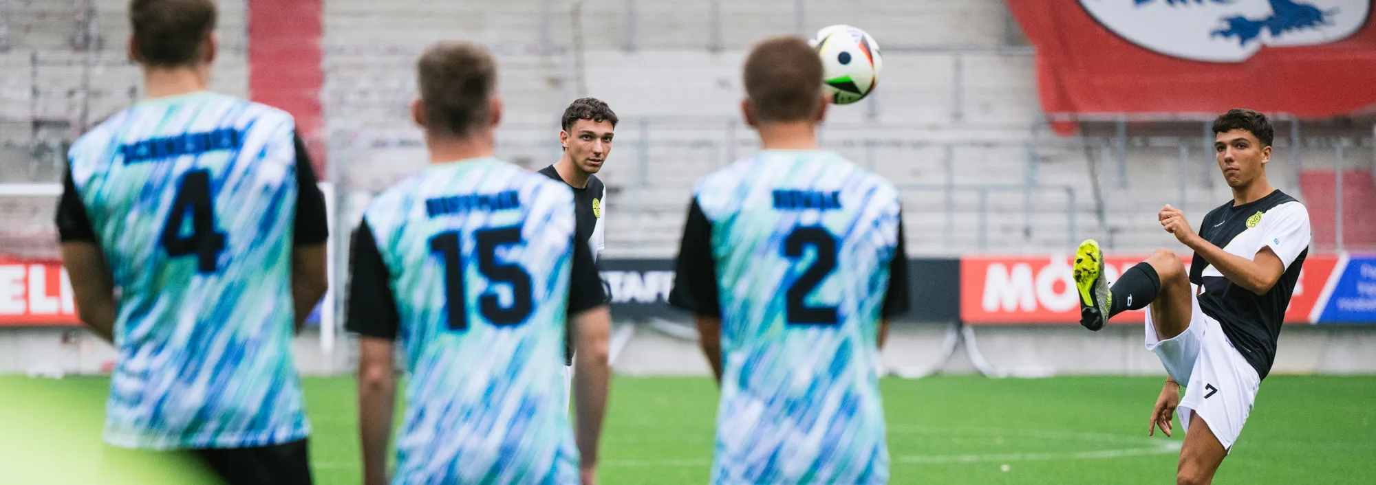 Un grupo de futbolistas con camisetas coloridas está en el campo, mientras uno de ellos levanta un balón con el pie.