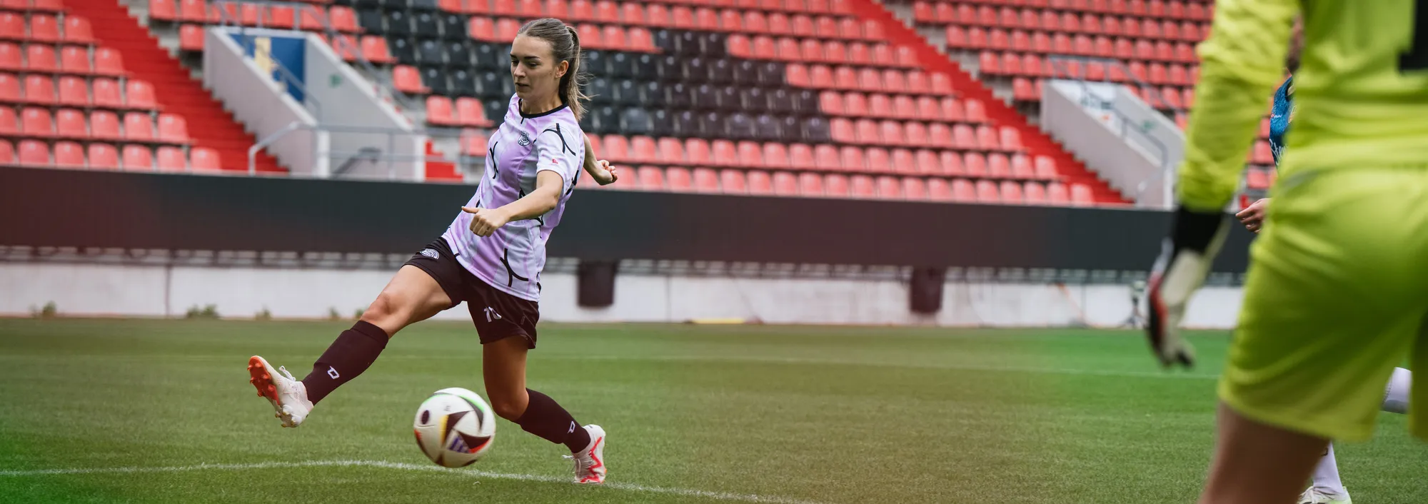 Una jugadora con una camiseta de fútbol morada está pateando el balón en un campo de césped.