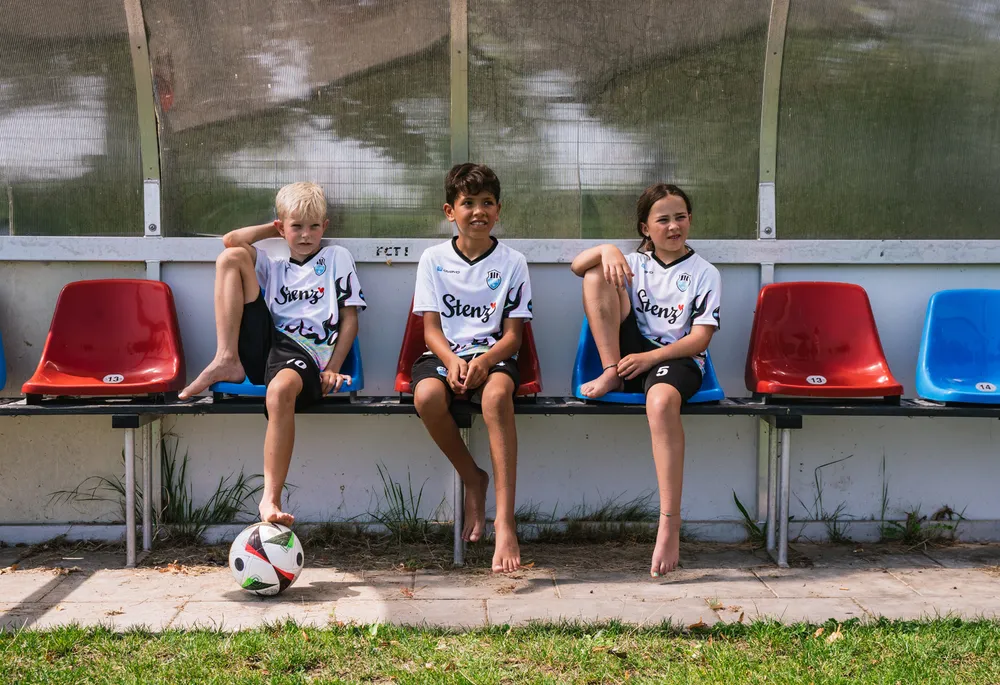 Tres niños en camisetas de fútbol deportivas están sentados en un banco junto a un balón de fútbol.
