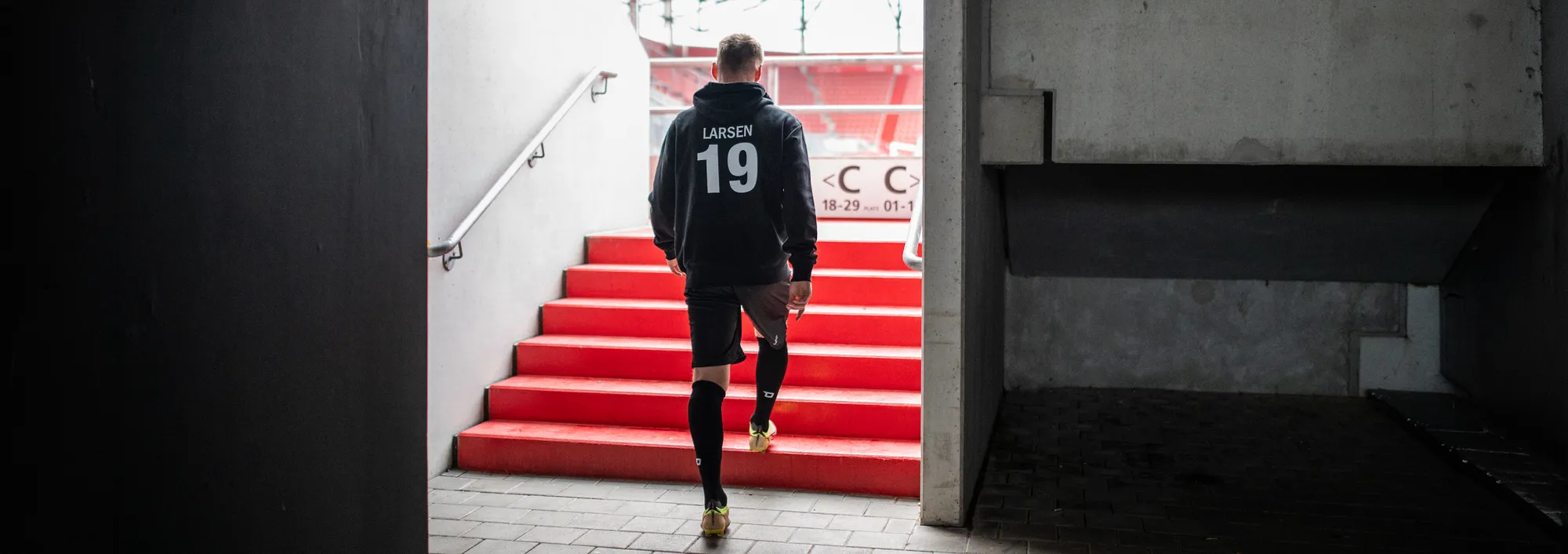 Un jugador con una sudadera oscura y pantalones cortos sube las escaleras rojas de un estadio.