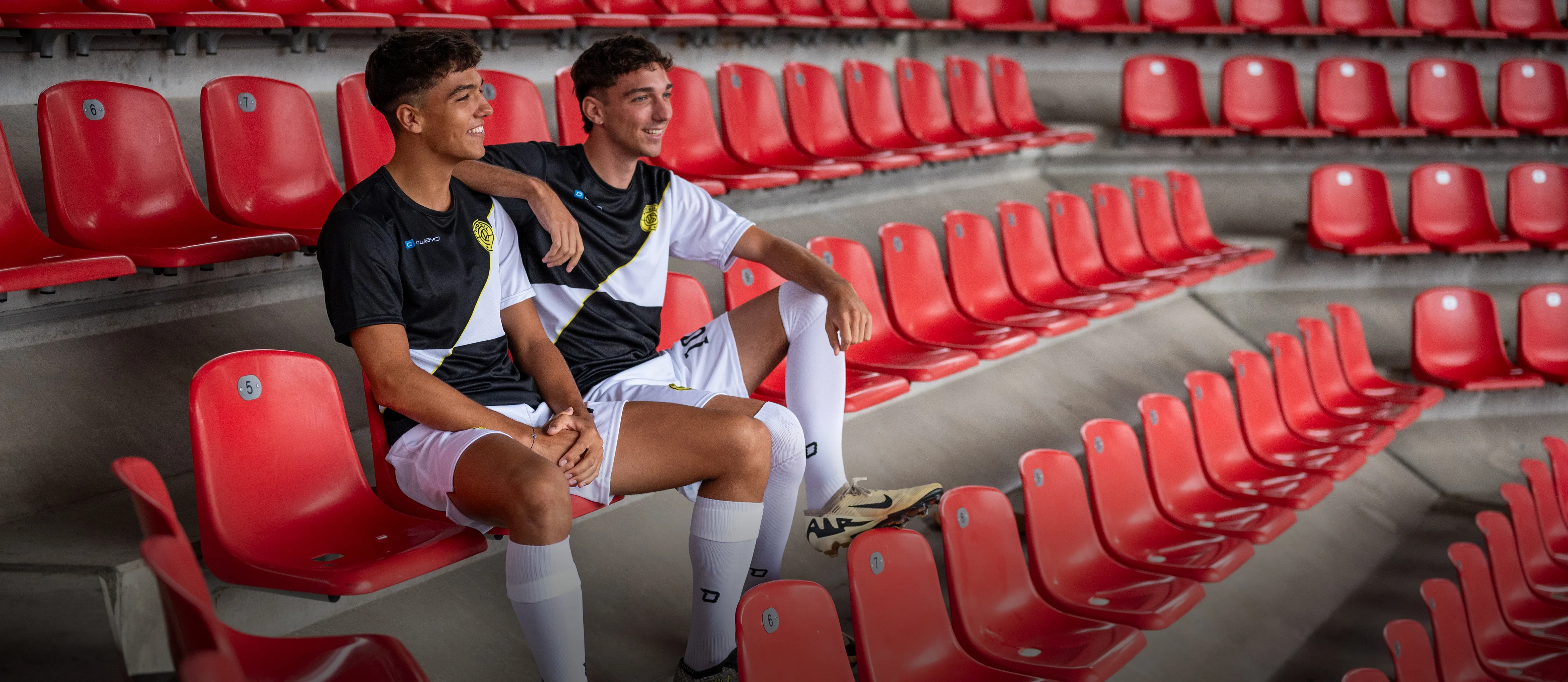 Dos jóvenes en ropa deportiva de fútbol están sentados de manera casual en asientos rojos de estadio.