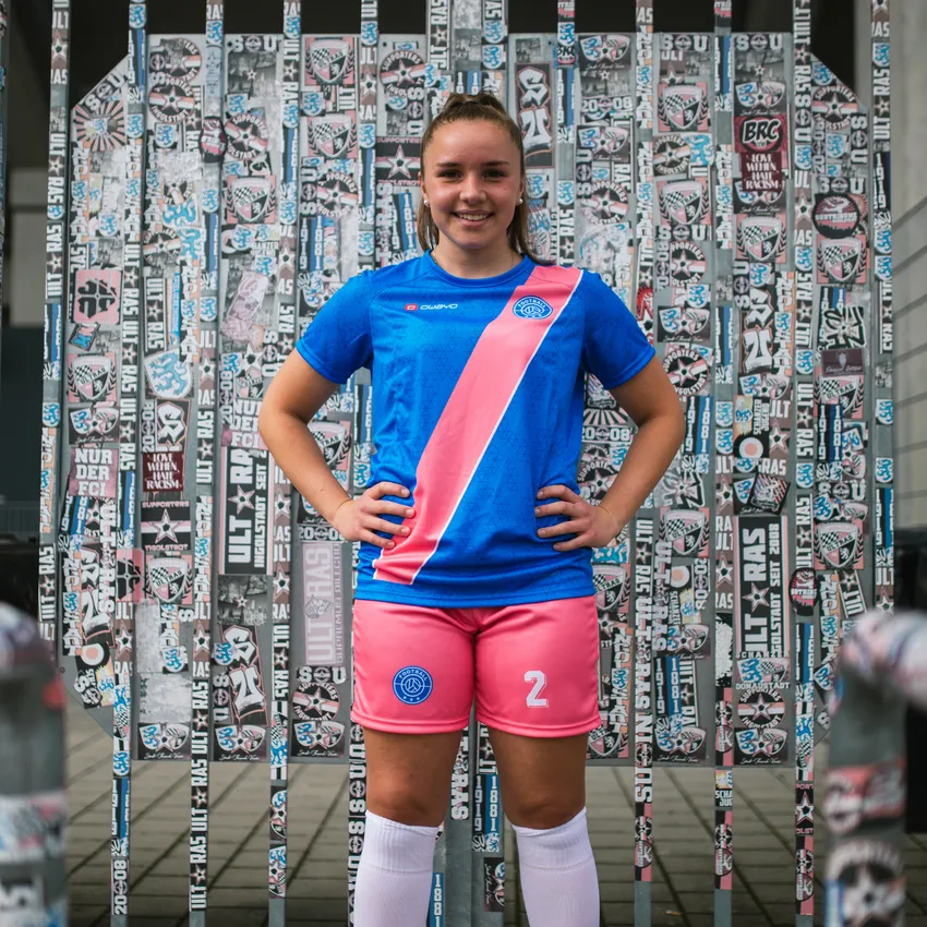 Una niña joven con una camiseta de fútbol azul con una franja rosa y pantalones cortos rosas se encuentra frente a una pared cubierta de pegatinas.