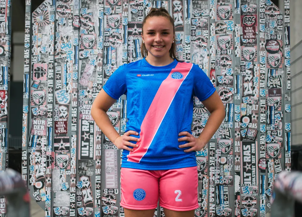 Una joven atleta posa con una camiseta de fútbol azul con una franja rosa y pantalones cortos rosas frente a una pared colorida cubierta de pegatinas.