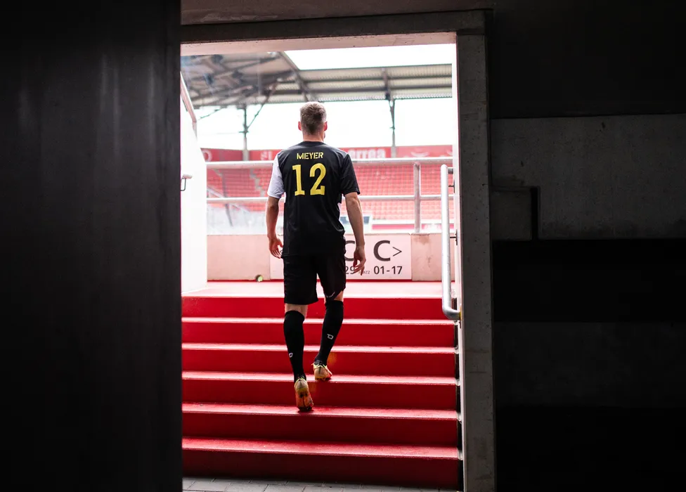 Un futbolista con una camiseta negra con el número 12 sube las escaleras rojas del estadio.