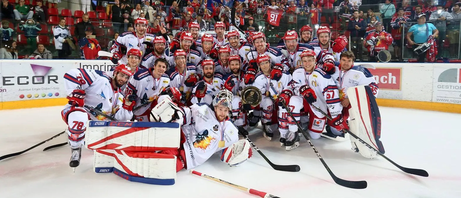 Un equipo de hockey sobre hielo con uniformes blancos con detalles rojos y azules posa en la pista, rodeado de aficionados, mientras los jugadores llevan medallas y sostienen un trofeo.