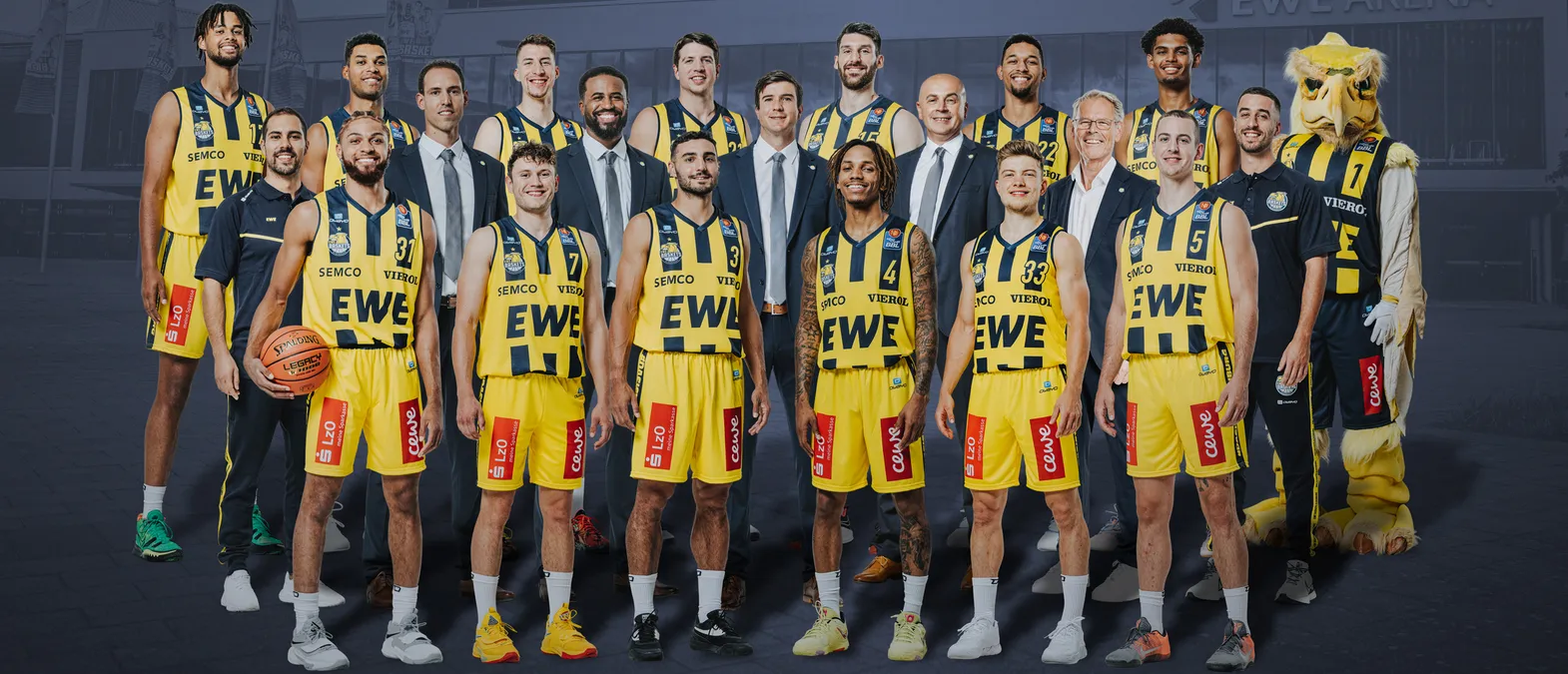 Una foto de grupo de un equipo de baloncesto con uniformes amarillos y negros con los patrocinadores 'EWE' y 'SEMC', una mascota y un balón de baloncesto, frente a un edificio.