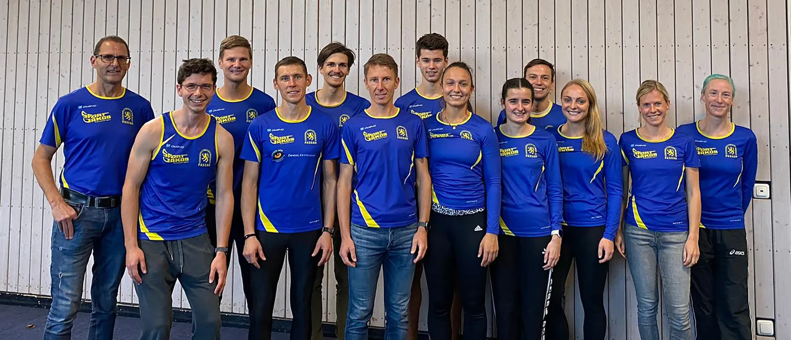Un grupo de hombres y mujeres con ropa deportiva azul y amarilla con el logo 'Sport Jakob Passau' posan frente a una pared de madera clara.