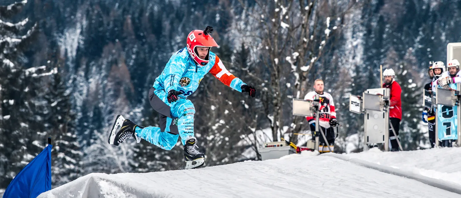 Un jugador de hockey sobre hielo con un jersey azul y blanco personalizado con logos de patrocinadores y un casco rojo salta sobre una rampa de nieve en una pista de hielo, con otros jugadores y árboles en el fondo.