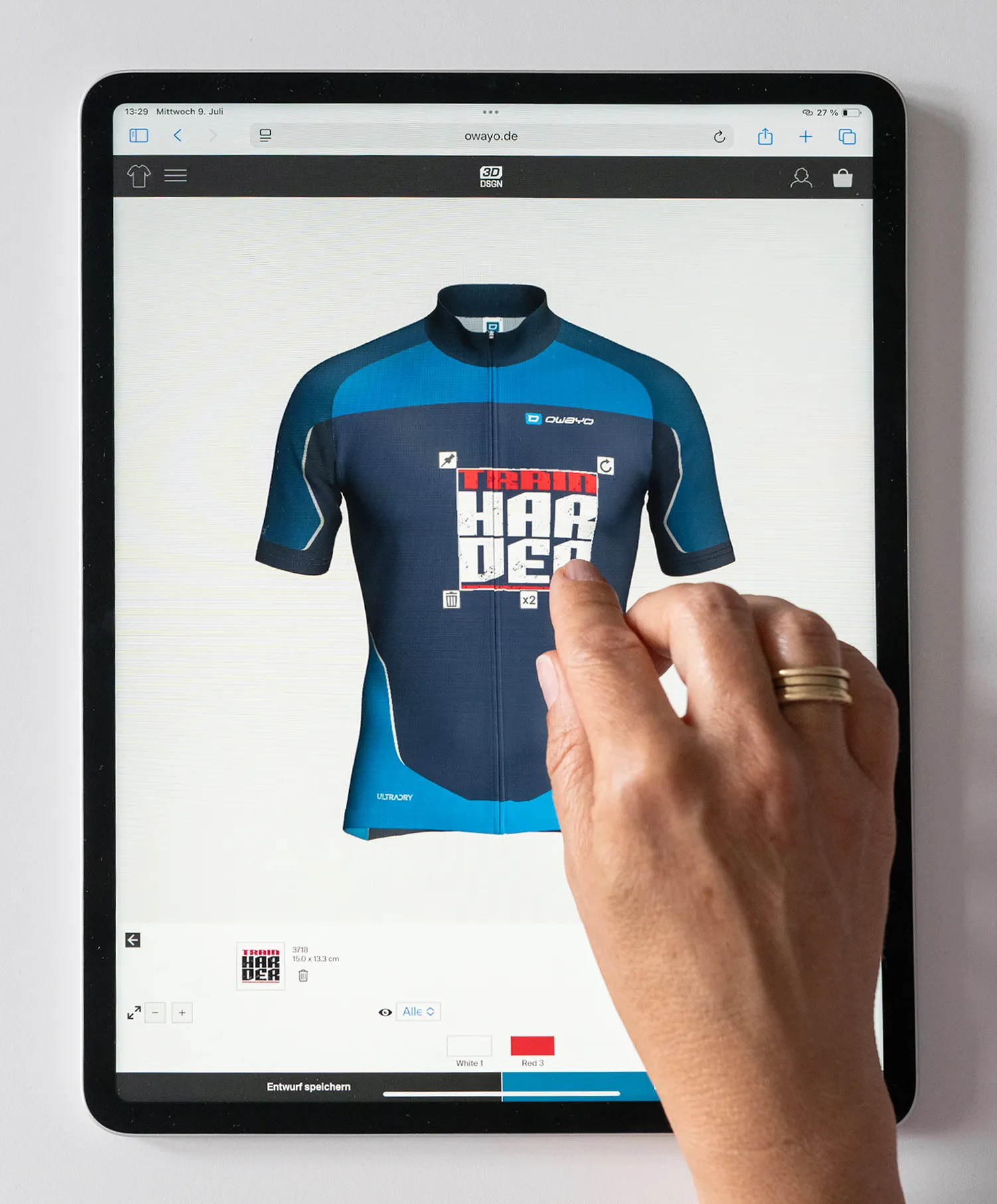 Una mano interactúa con una tableta que muestra el sitio web de owayo, mostrando una camiseta de ciclismo personalizada azul y azul oscuro con el texto 'TRAIN HARDER'.