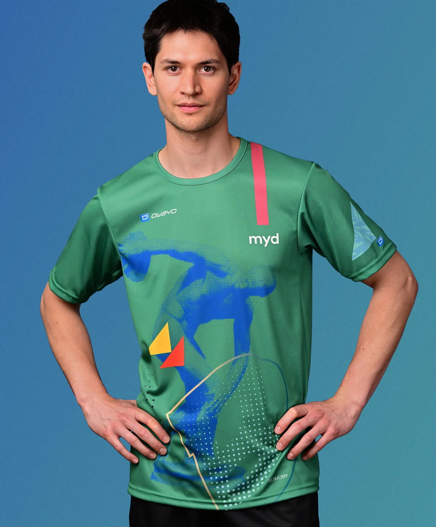 Un hombre con una camiseta deportiva verde personalizada con un diseño geométrico azul, amarillo y rojo que representa una figura deportiva, de pie frente a un fondo azul.